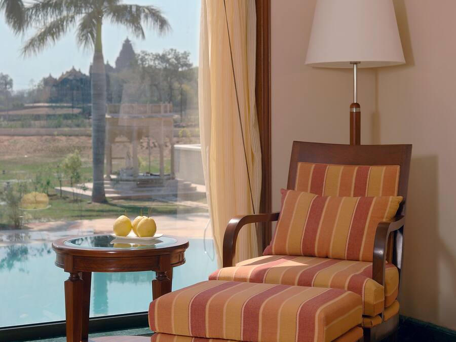 Una habitación con vistas a palmeras y un templo a través de una ventana, una silla de madera con cojines a rayas, una mesita redonda con dos limones y una lámpara de pie.