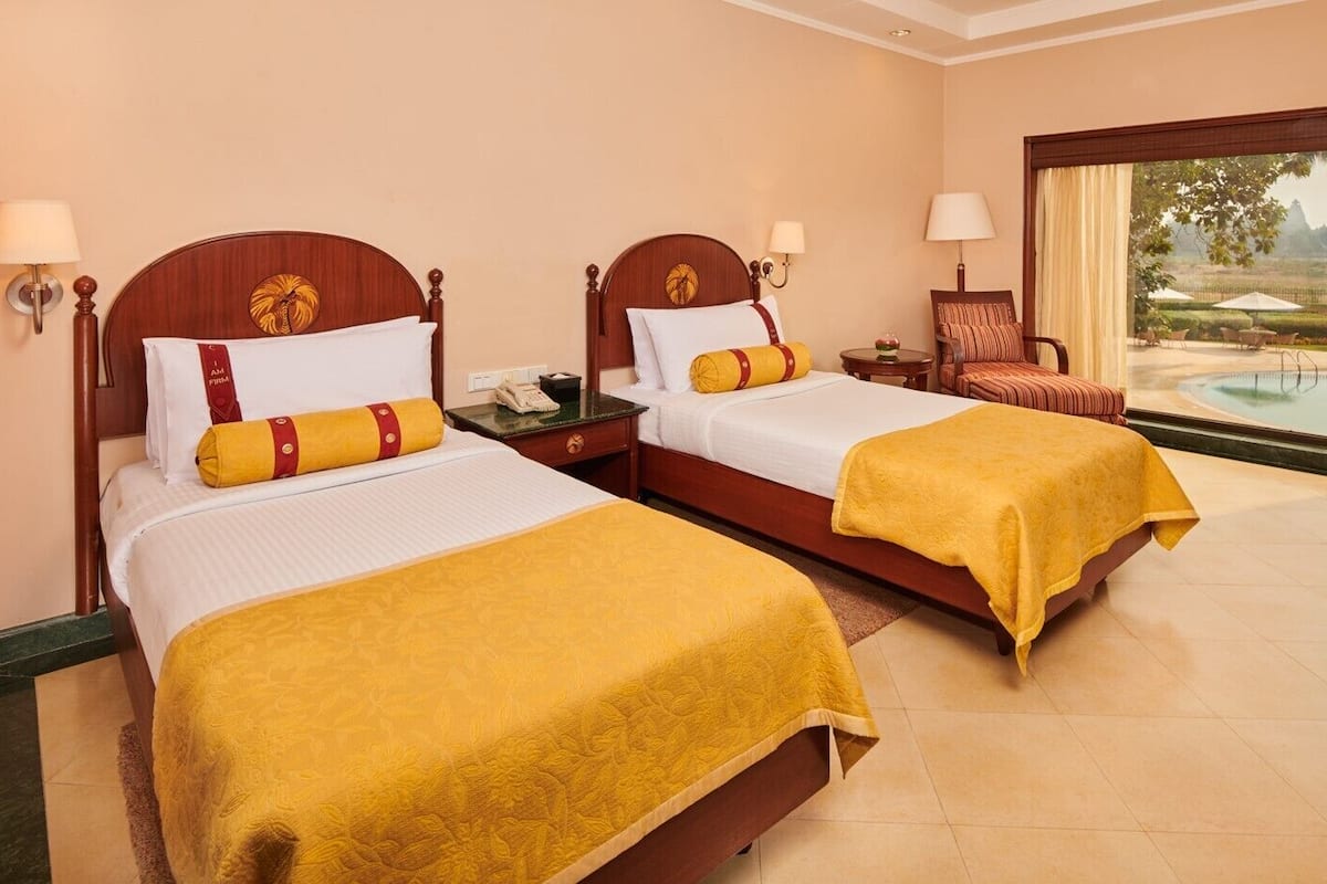 A hotel room with two beds, each with a wooden headboard, white bedding, and a yellow throw blanket. There is a small table with a lamp between the beds, and a chair with a cushion. A large window offers a view of a pool and landscape.