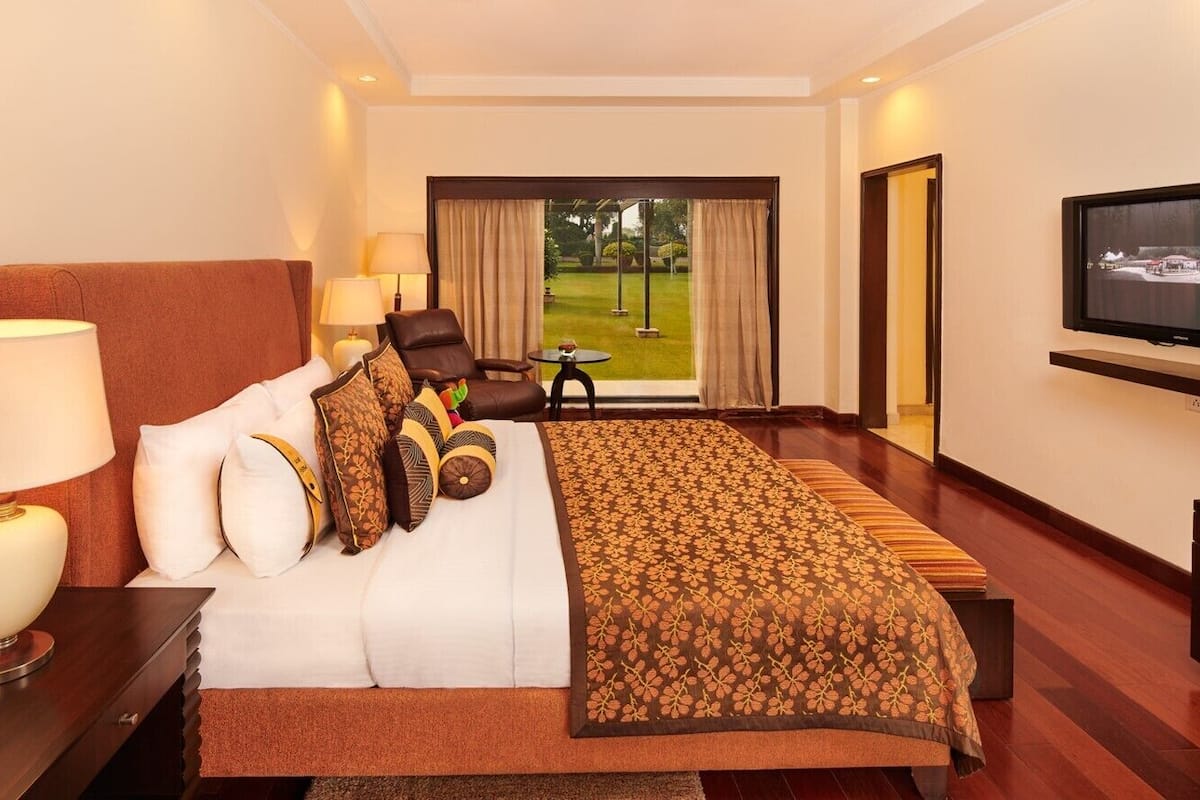 A hotel room with a large bed, a TV, a desk with a lamp, a chair, and a view of a garden through a window.