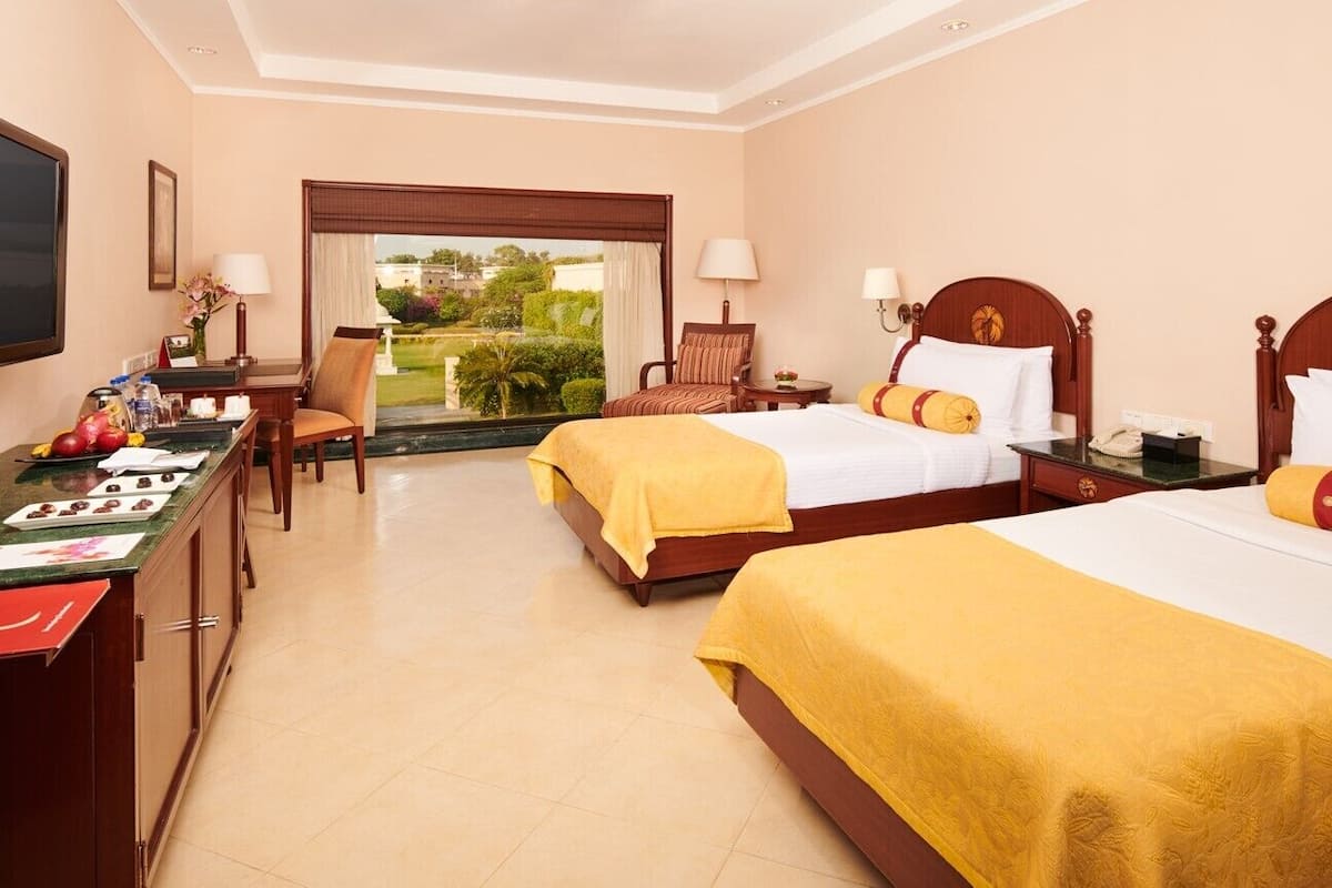 A hotel room with two beds, a desk with a flat-screen TV, a chair, and a view of the garden.