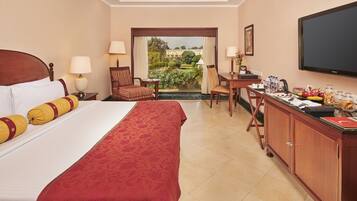 A hotel room with a large bed, a desk with a chair, a television, and a view of a garden through a window.