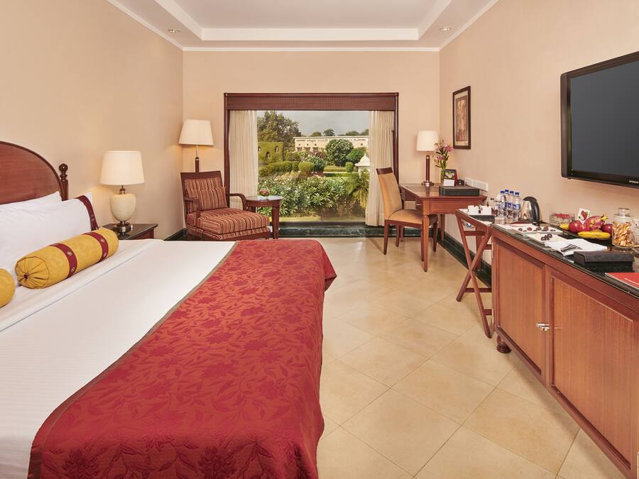 A hotel room with a large bed, a desk with a chair, a television, and a view of a garden through a window.