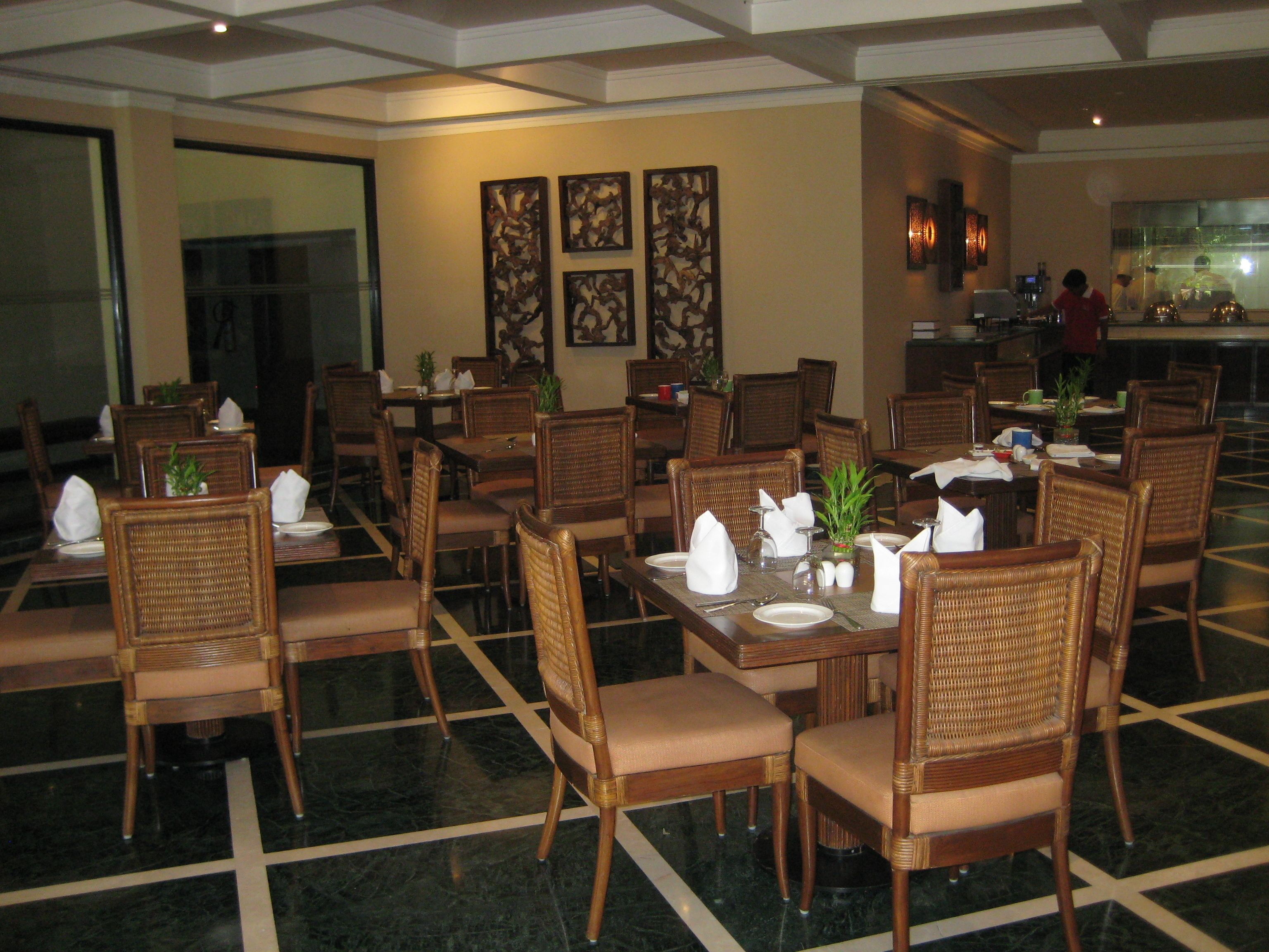 A dining area with wicker chairs and tables, some with napkins and cutlery. There are wall art pieces and a kitchen area in the background.