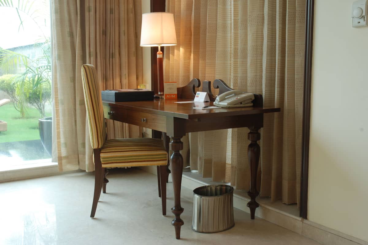A hotel room with a wooden desk, a striped chair, a lamp, a telephone, and a trash can.