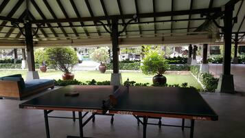 An outdoor table tennis table with a canopy overhead, surrounded by greenery and potted plants.