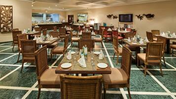A hotel dining area with multiple tables set for guests, featuring wicker chairs and a marble floor.