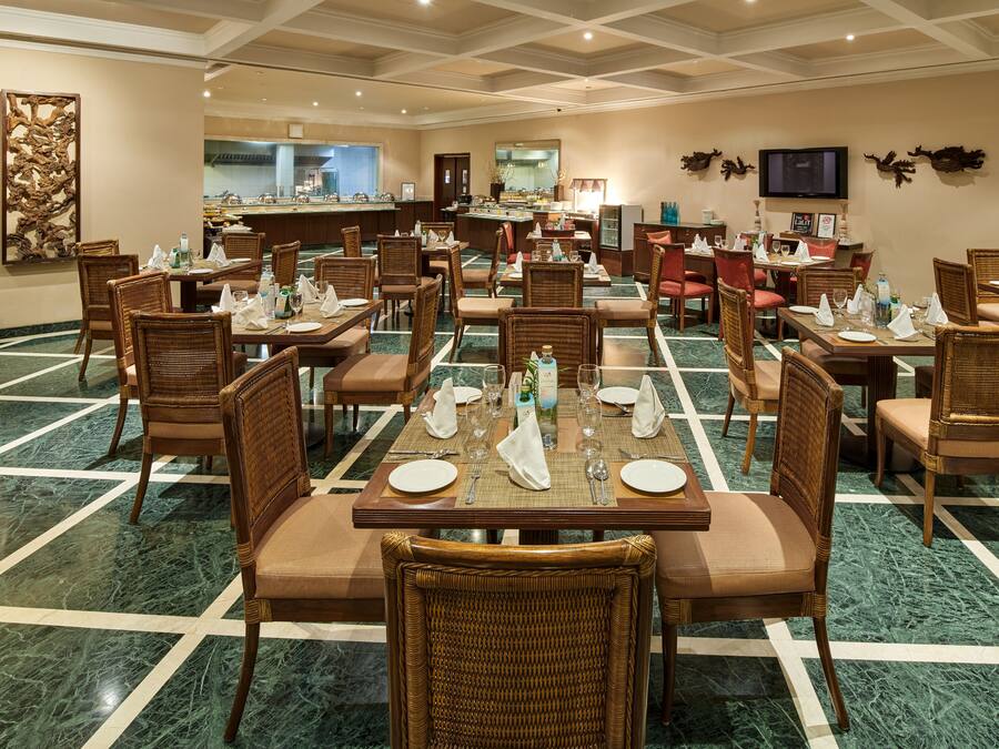 A hotel dining area with multiple tables set for guests, featuring wicker chairs and a marble floor.