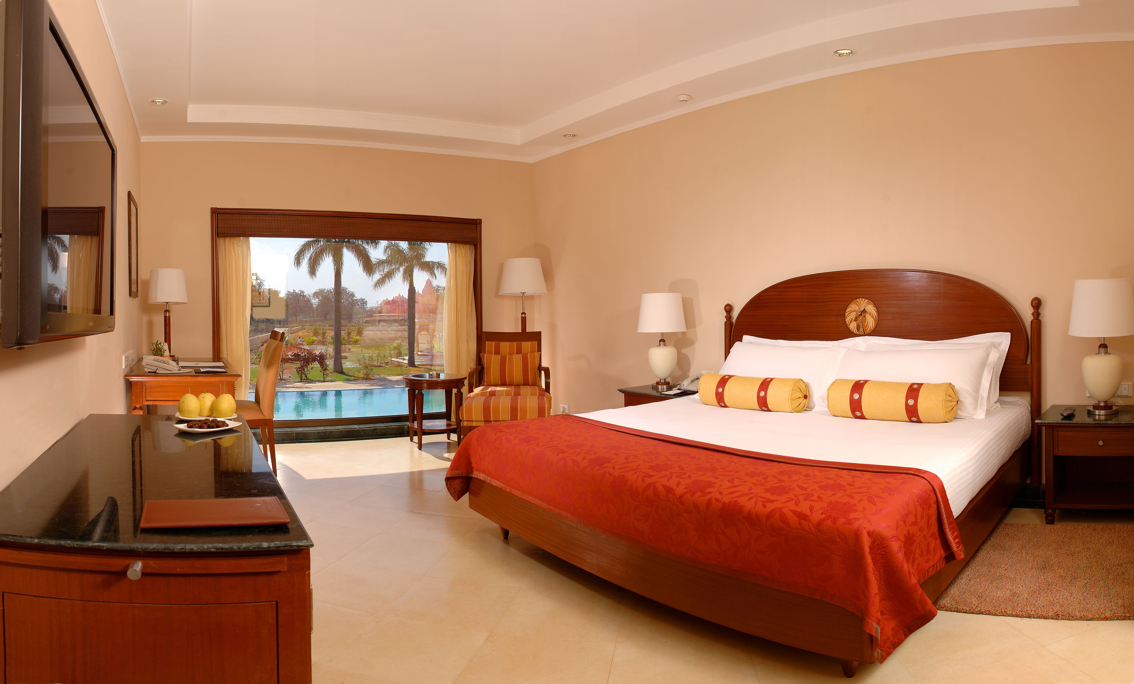 A hotel room with a large bed, two bedside tables with lamps, a desk with a chair, and a view of palm trees and a pool.