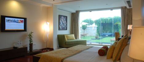 A hotel room with a bed, a green armchair, a ceiling fan, a TV on the wall, and a large window with a view of the outdoors.