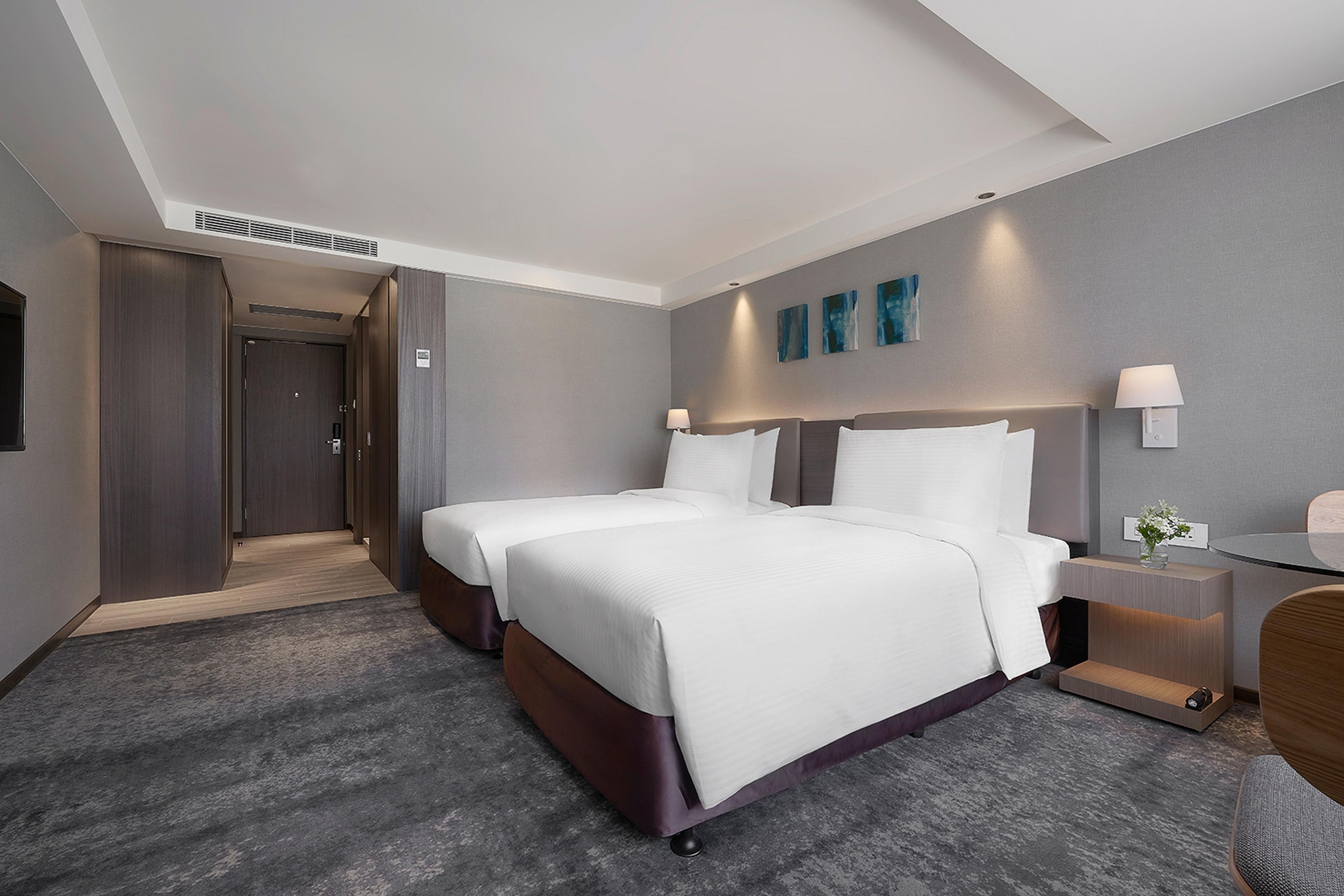 Habitación de hotel con dos camas, un escritorio, una silla, una mesita de noche y una lámpara de pared.