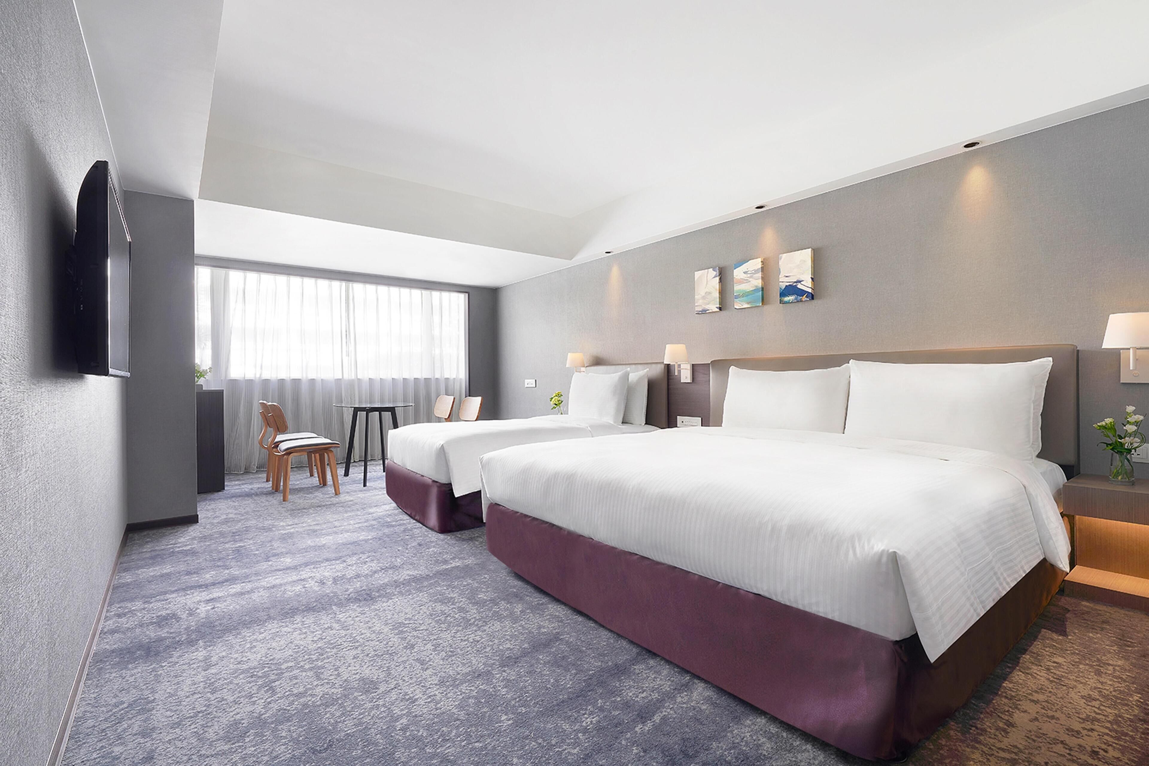 A hotel room with two beds, a desk, a chair, and a large window.