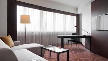 A hotel room with a sofa, a small table, a desk, a chair, a lamp, and a large window with sheer curtains.