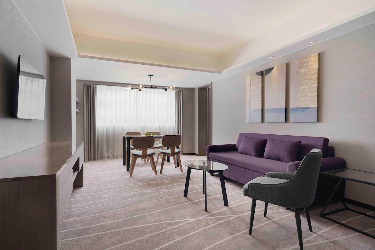 A modern hotel room with a sofa, a dining table with chairs, a TV mounted on the wall, and a large window with curtains.