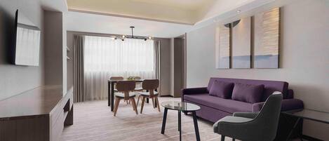 A modern hotel room with a sofa, a dining table with chairs, a TV mounted on the wall, and a large window with curtains.
