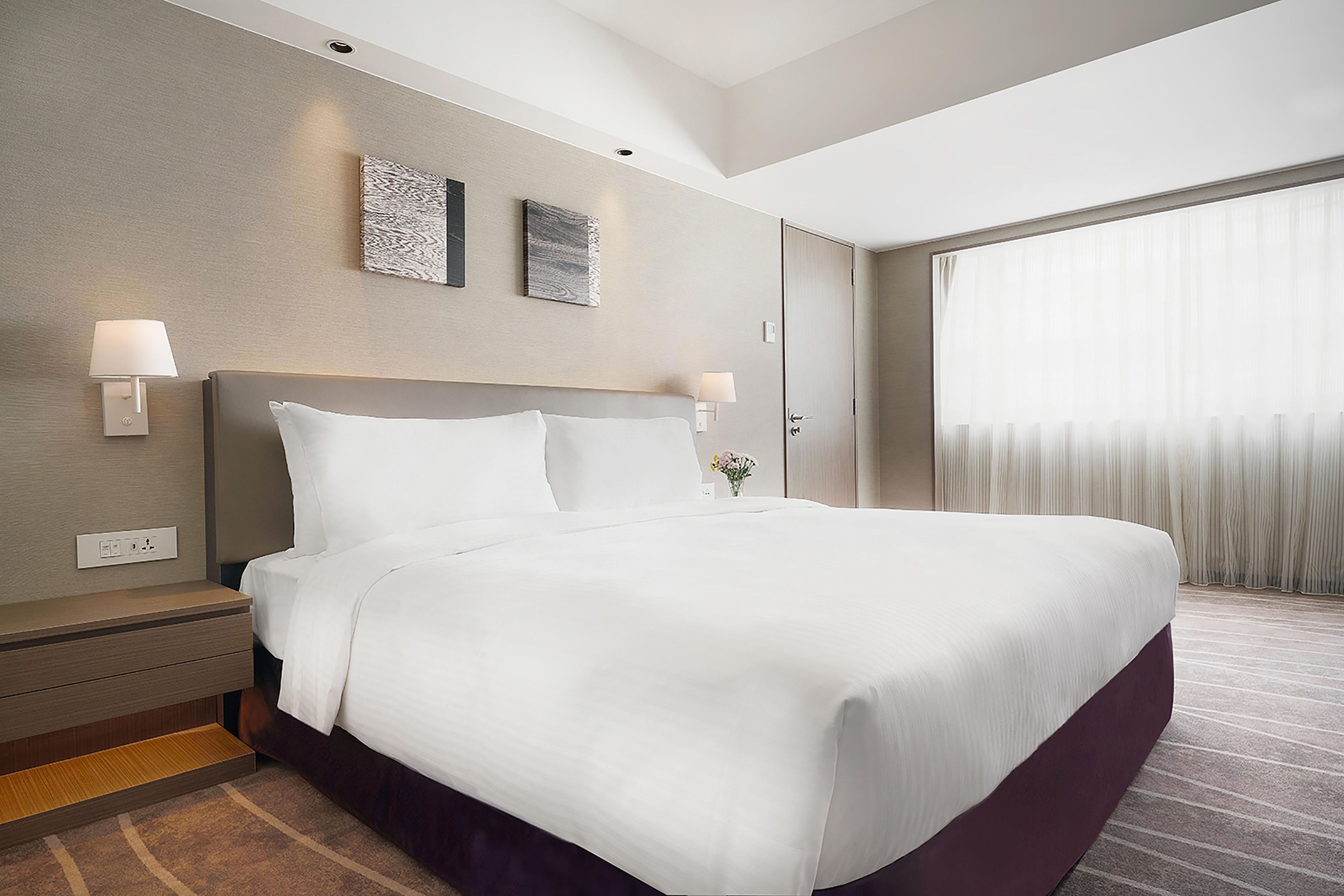 A hotel room with a large bed, two wall-mounted lamps, a nightstand, and a window with curtains.
