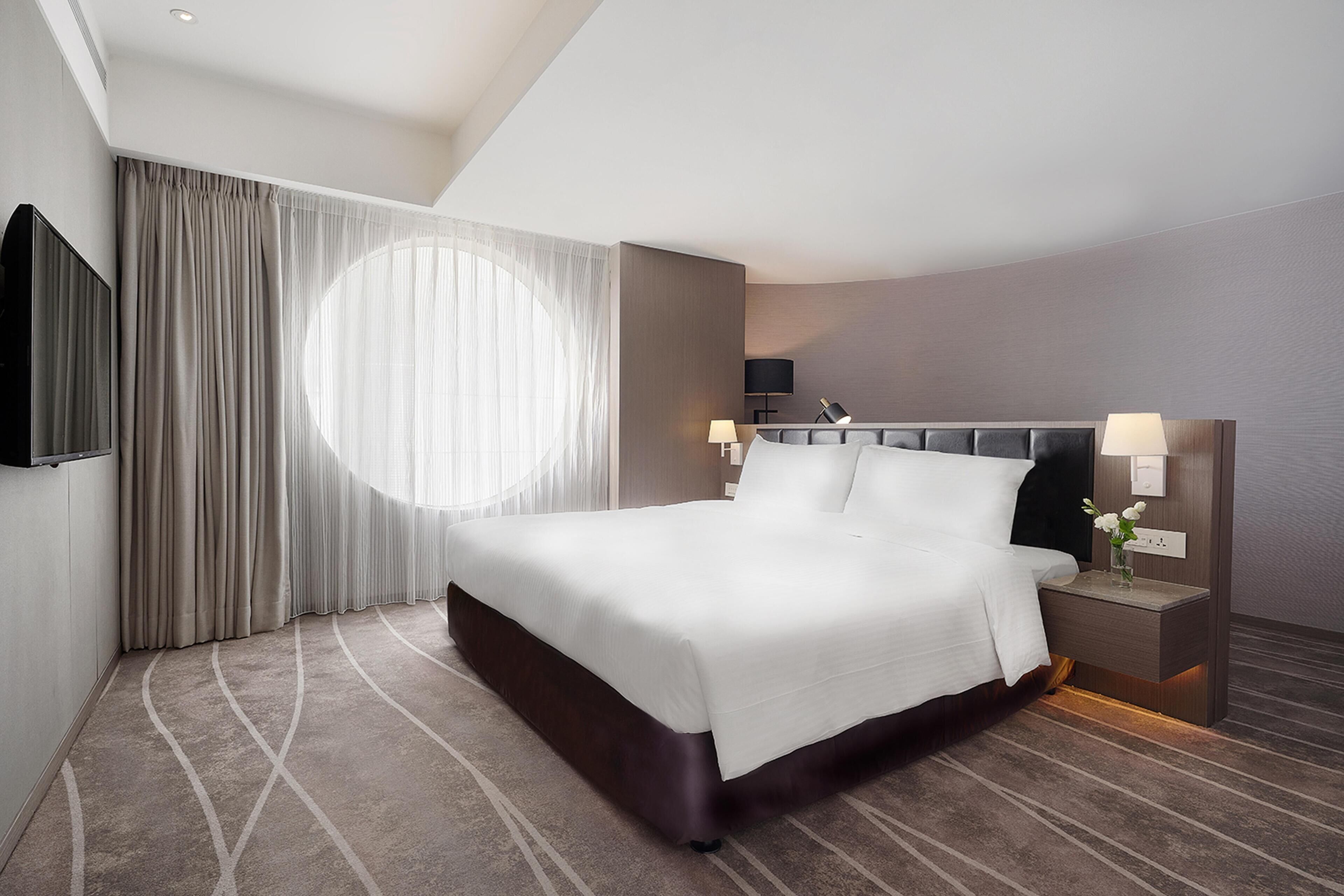 A hotel room with a large bed, a TV, a bedside table with a lamp, and a view of the outdoors through a window.