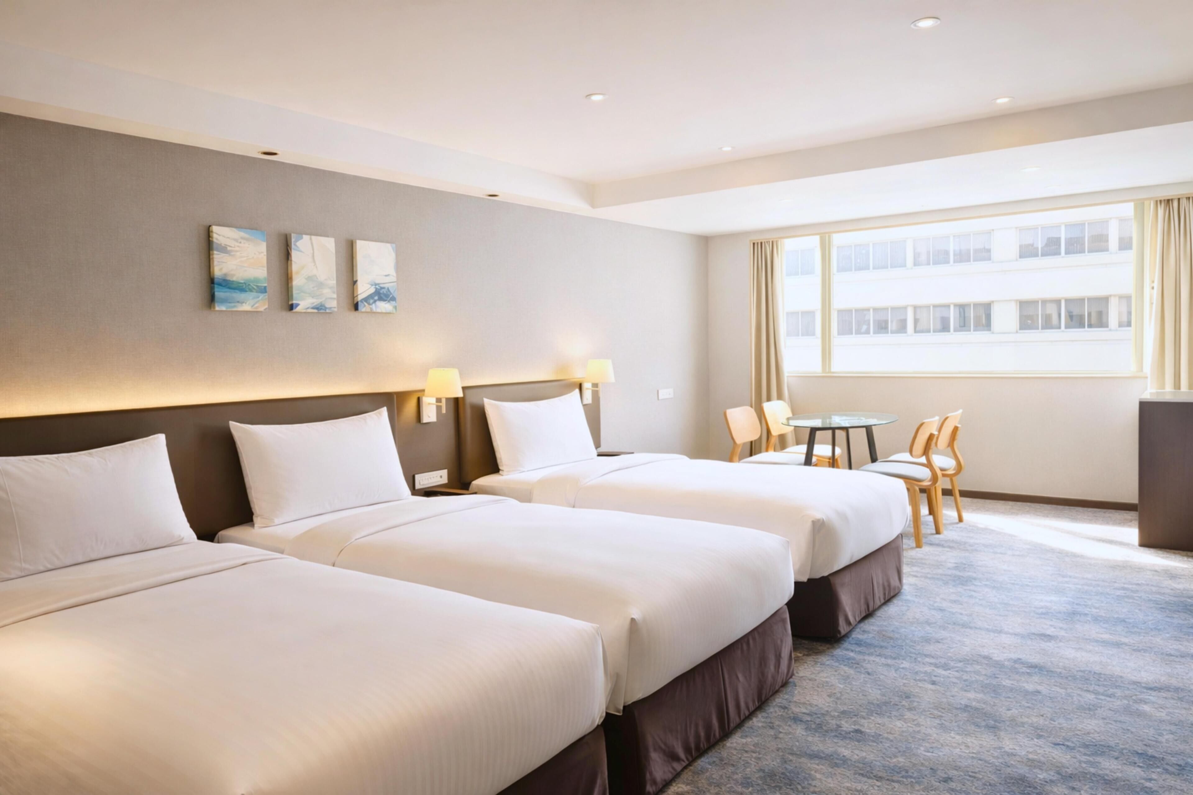 Une chambre d’hôtel avec trois lits, une petite table, des chaises et une grande fenêtre.