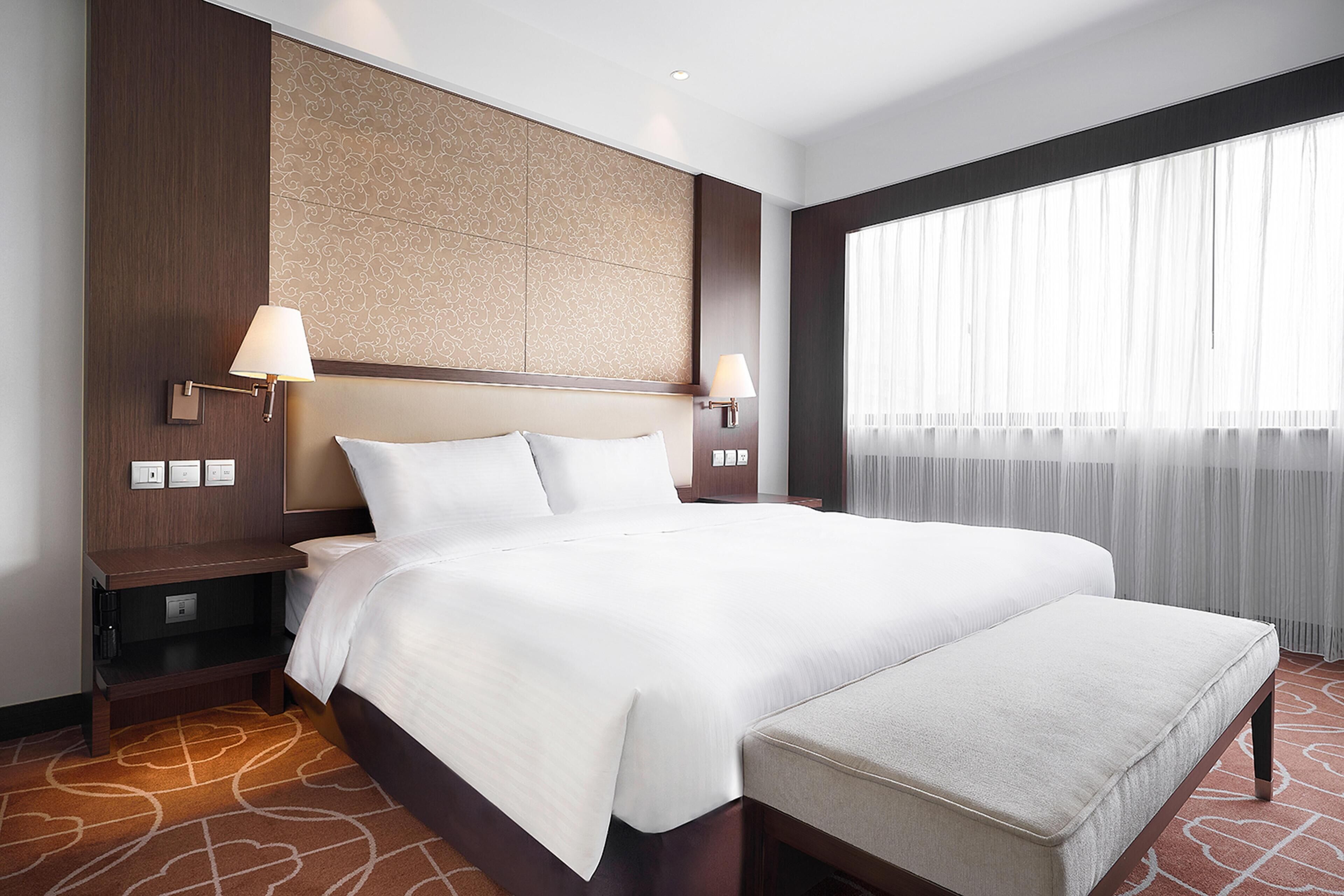 A large bed with a headboard, two bedside lamps, a nightstand, and a window with sheer curtains.