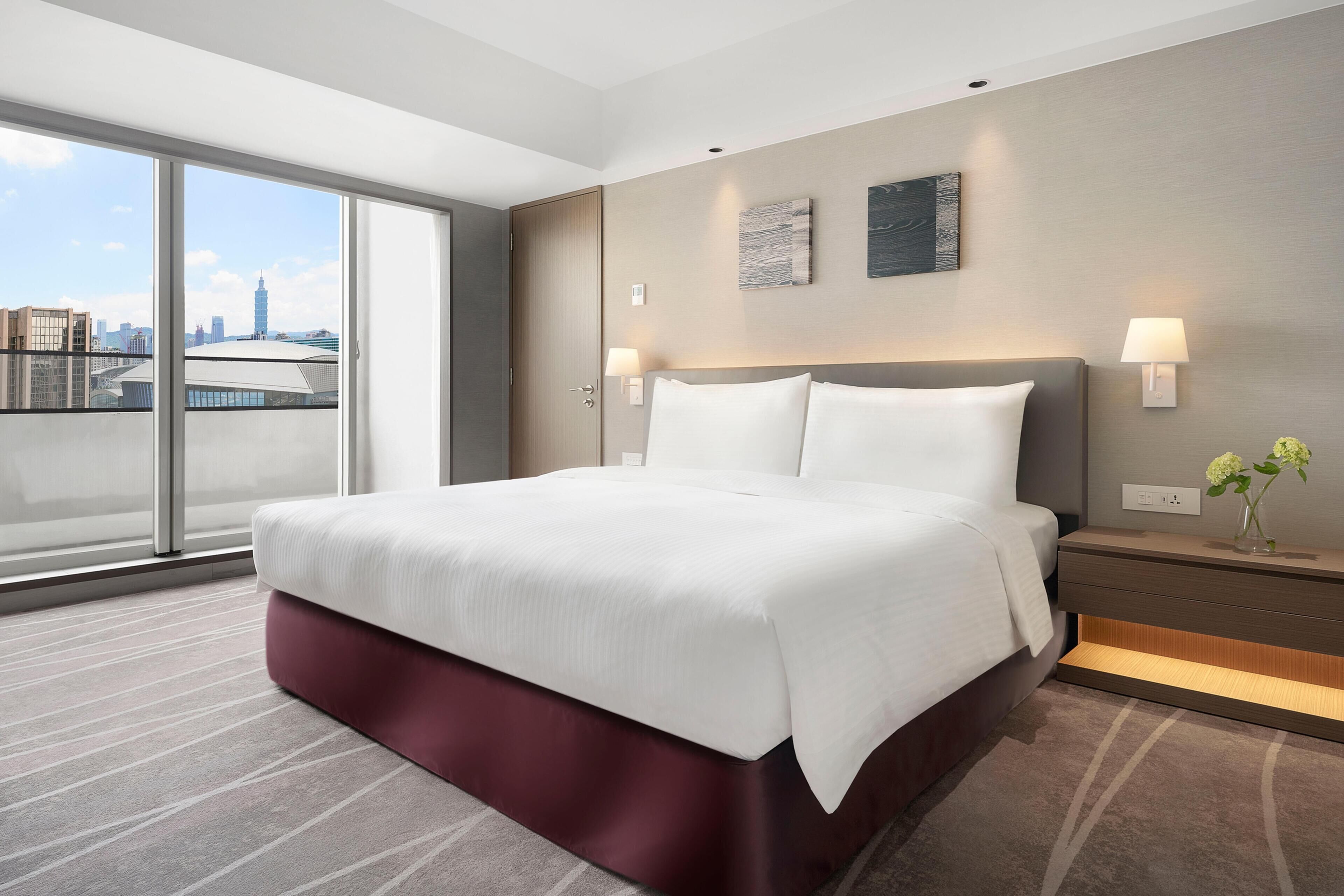 Een moderne hotelkamer met een groot bed, een nachtkastje, een lamp en uitzicht op een stadsgezicht.