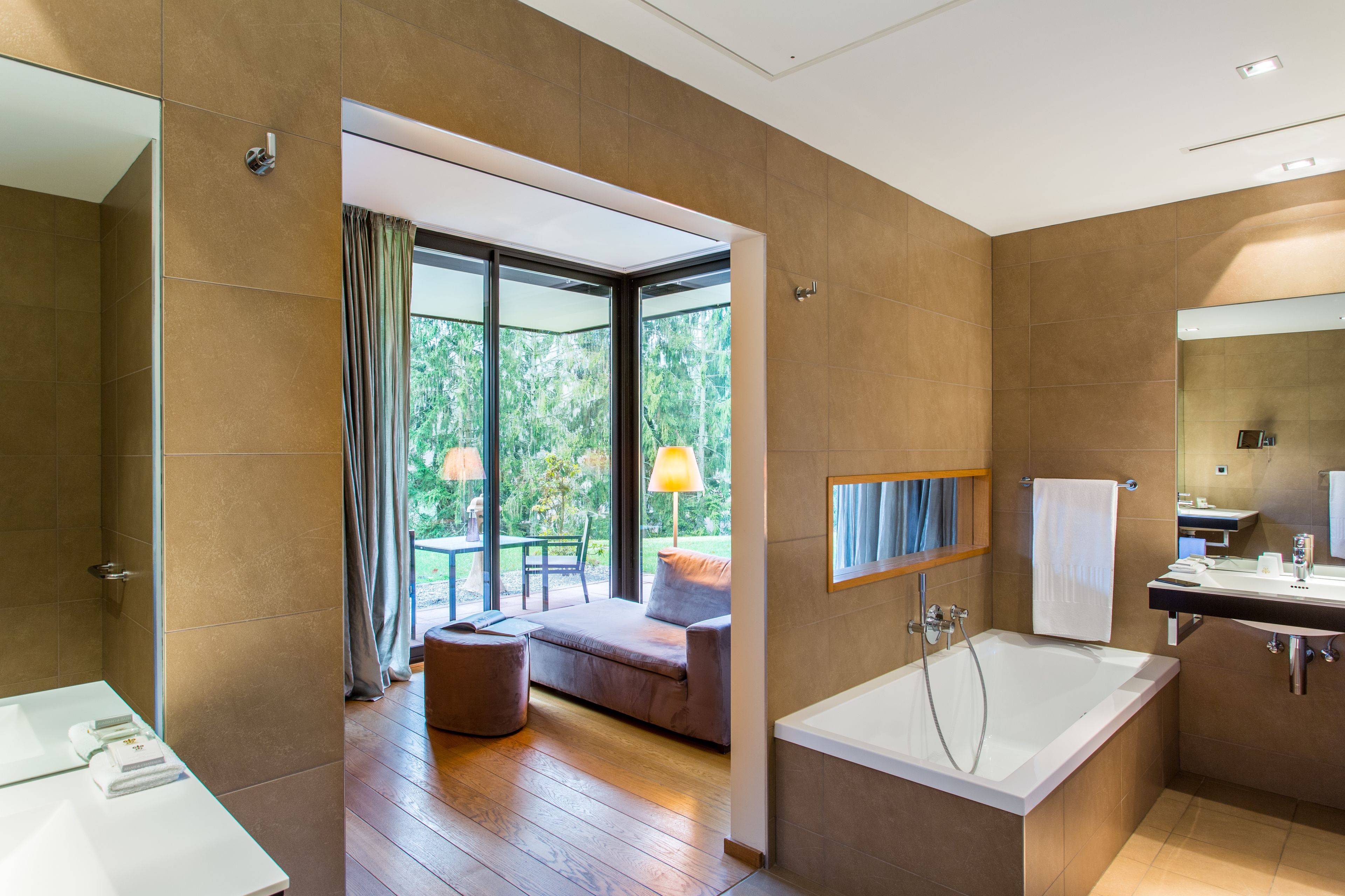 A modern bathroom with a large bathtub, a glass-enclosed shower area, and a seating area with a view of greenery.