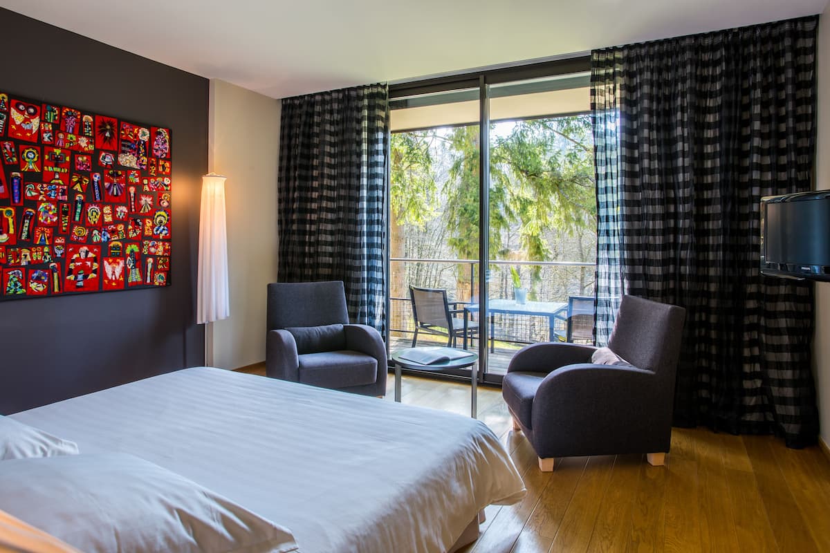 Una camera d'albergo con un letto grande, due poltrone, un tavolino, una TV e un balcone con vista sugli alberi.