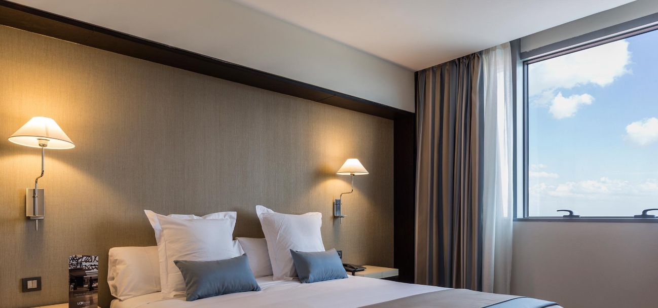 A hotel room with a bed, two bedside lamps, a window with curtains, and a view of the sky.