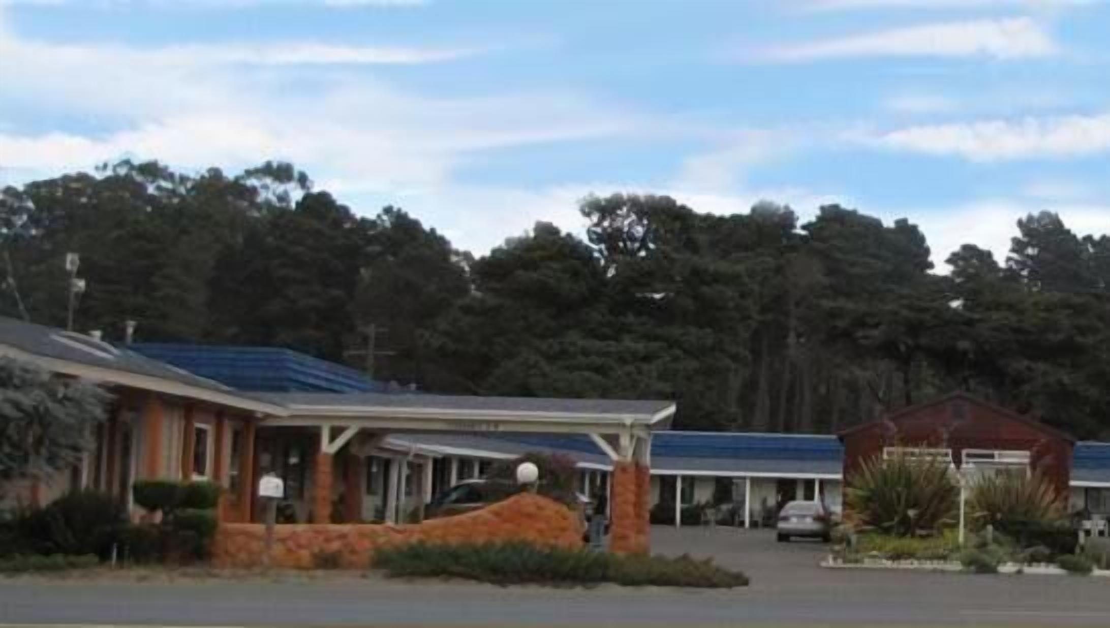A motel with a covered parking area, a small building with a red roof, and a large tree-lined background.
