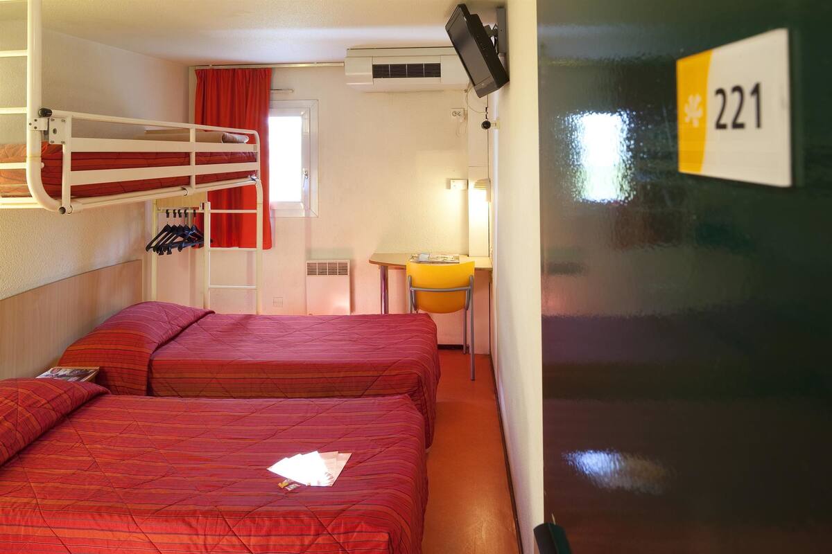A room with two beds, a small desk, a chair, and a window with red curtains.