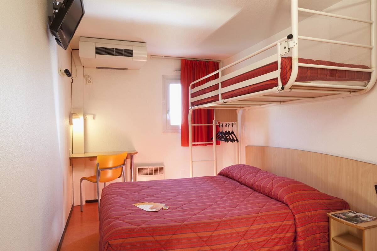A bunk bed with a red quilt, a desk with a chair, a TV mounted on the wall, and a window with red curtains.