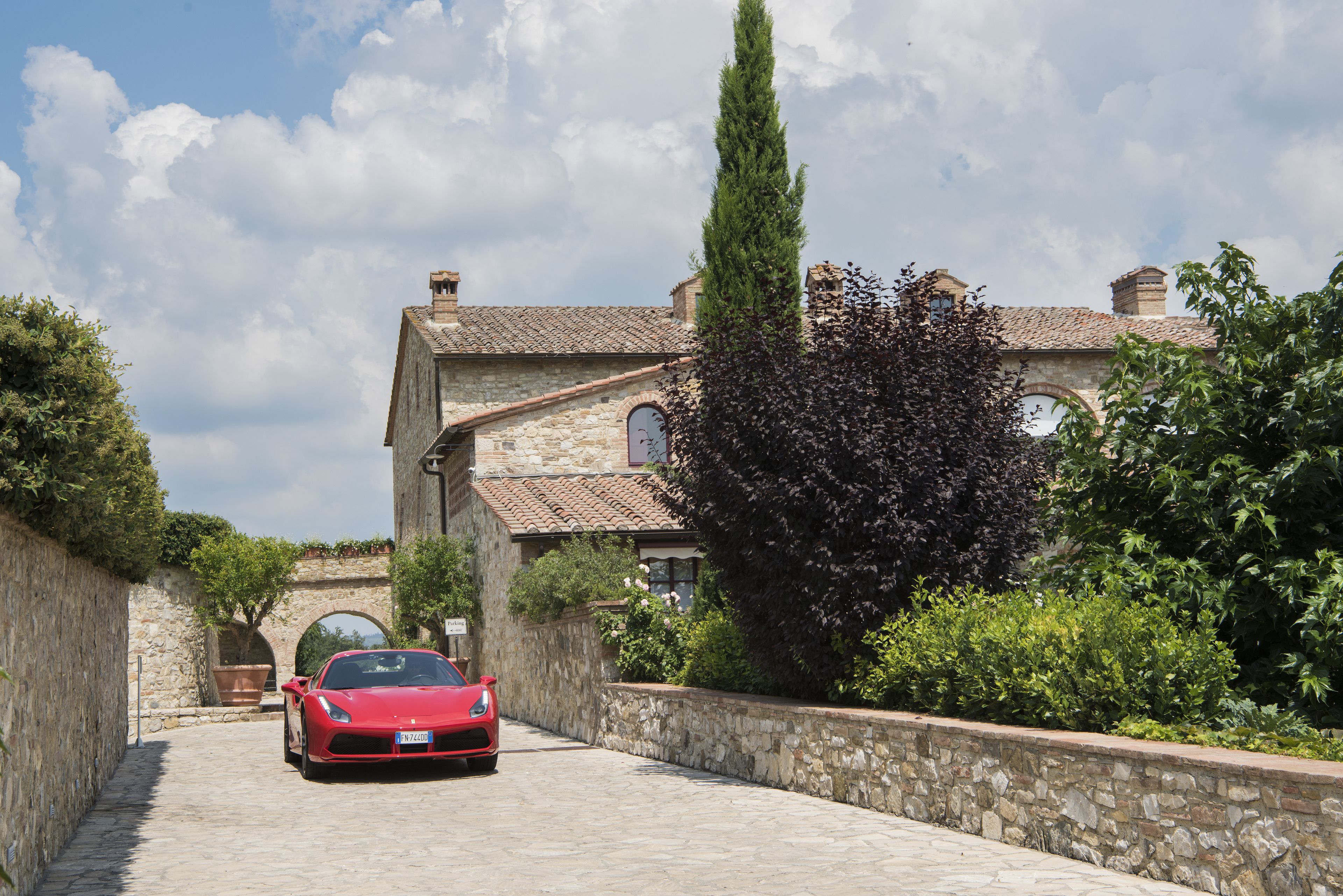 Une voiture de sport rouge est garée devant un bâtiment en pierre, doté d’un toit en tuiles et de fenêtres en arc de cercle.