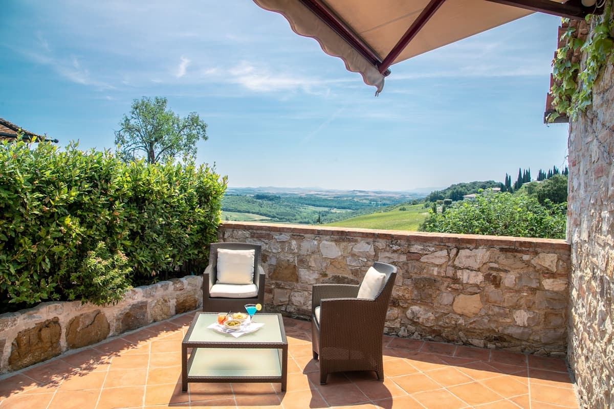 Una terraza con un muro de piedra, dos sillas de mimbre, una mesa con bebidas y aperitivos, y vistas al campo.