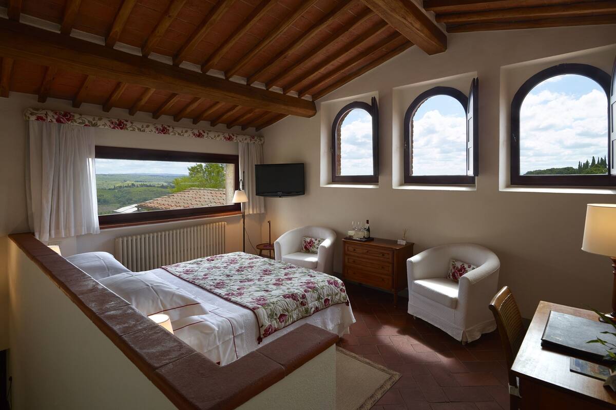 Un dormitorio con cama, dos sillas, cómoda, televisor y tres ventanas con vistas al campo.