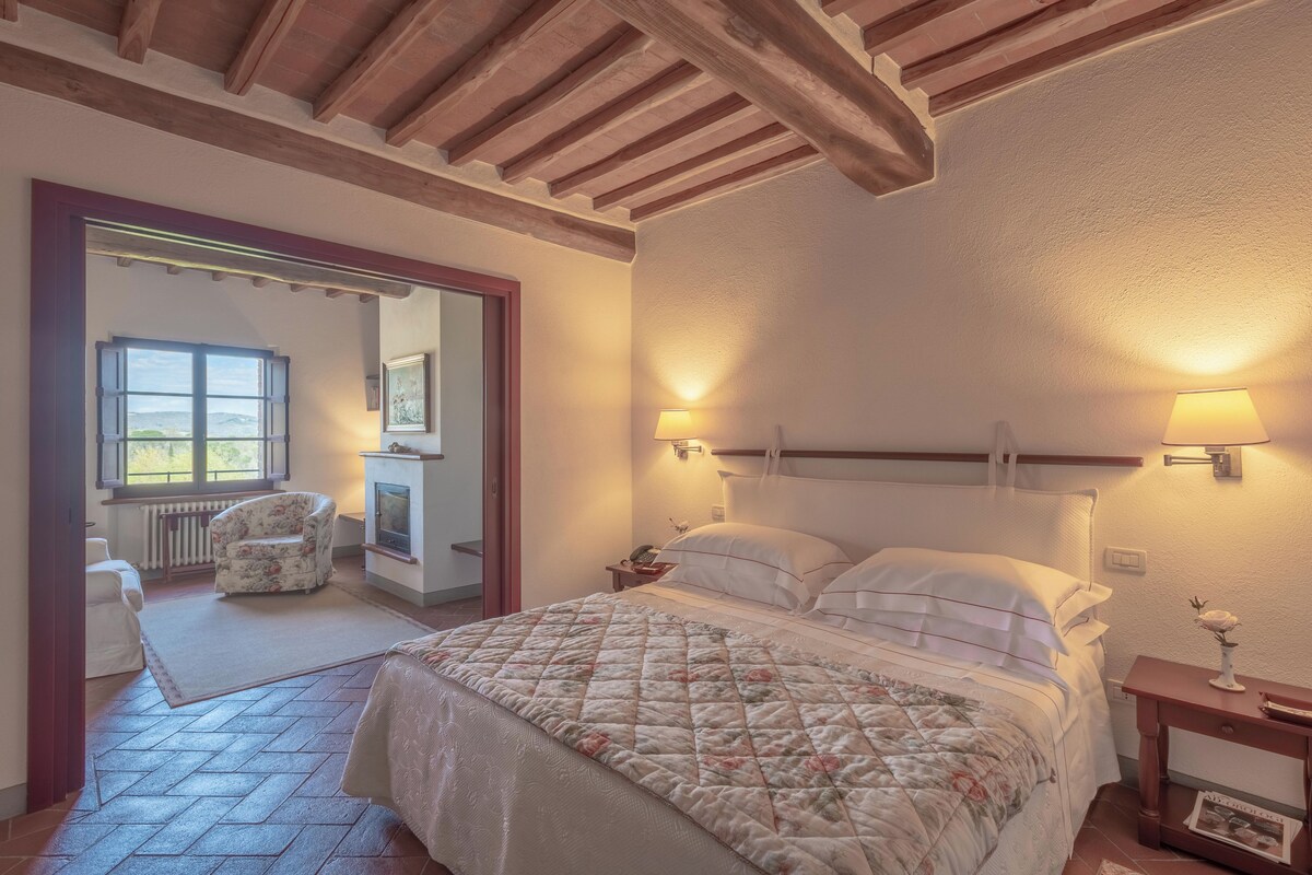 Une chambre avec un grand lit, une table de chevet, une lampe, une chaise et une vue sur une pièce avec une cheminée.