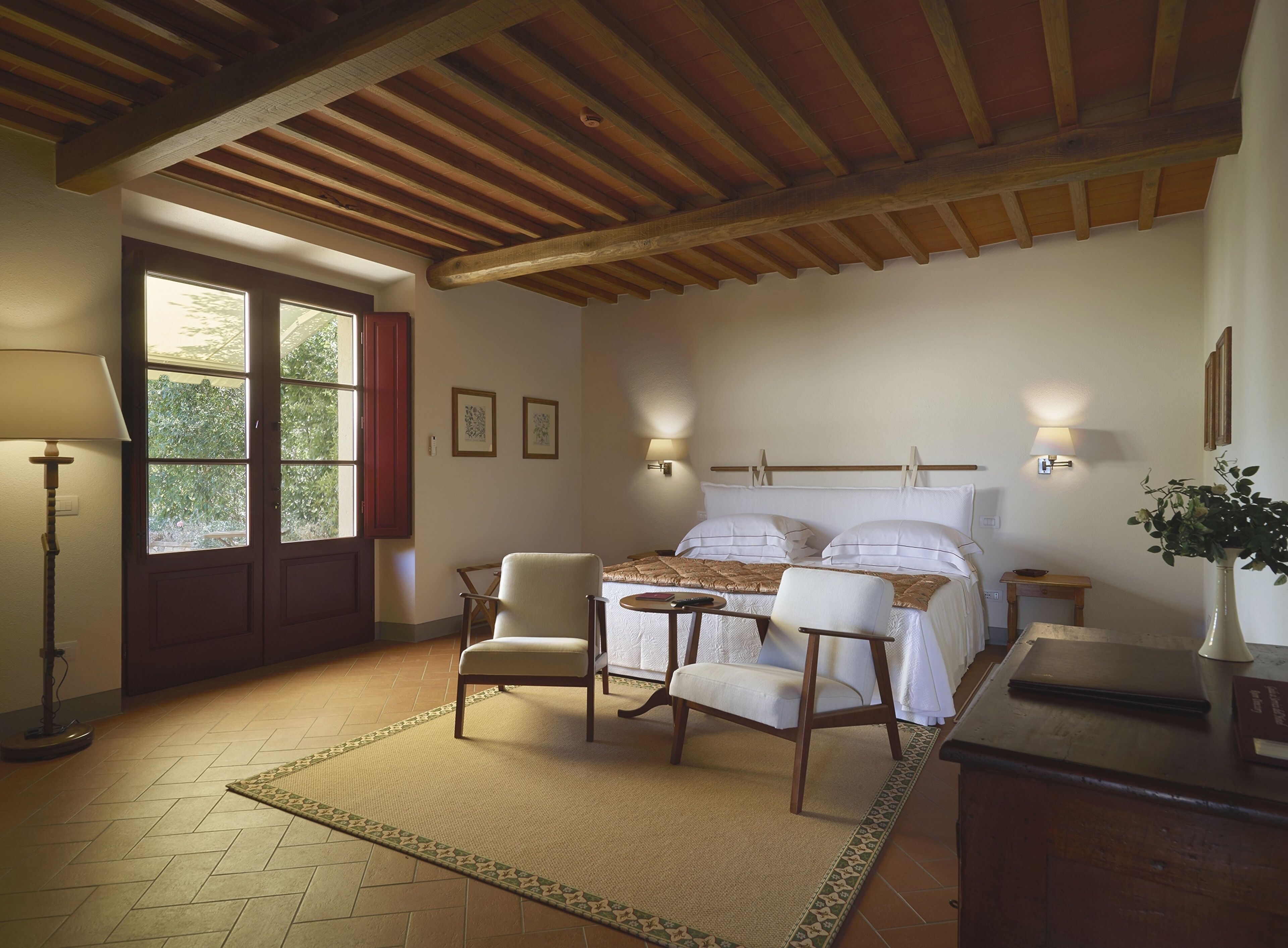 A bedroom with a bed, two chairs, a bedside table, a lamp, and a window with curtains.