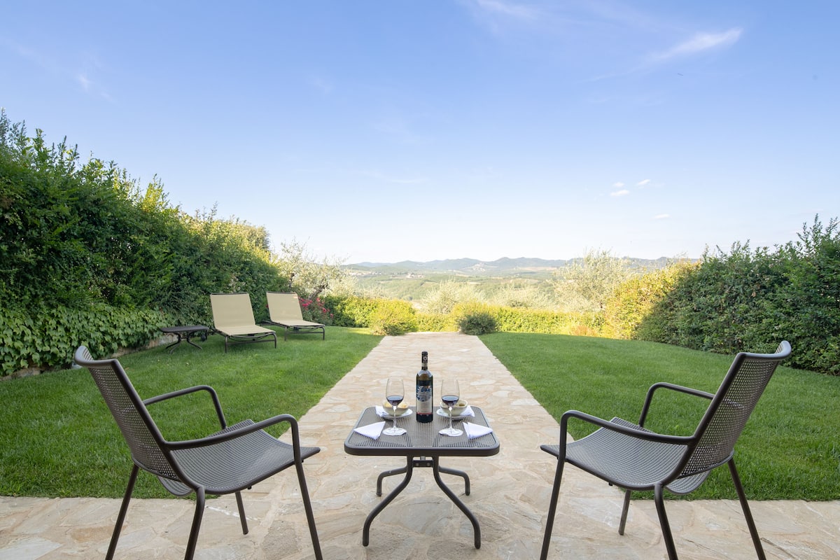 Un patio con una mesa lista para dos personas, dos sillas y una botella de vino.