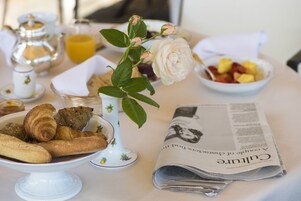 Una mesa de desayuno con cruasanes, un florero con una rosa blanca, un periódico y un bol de fruta.