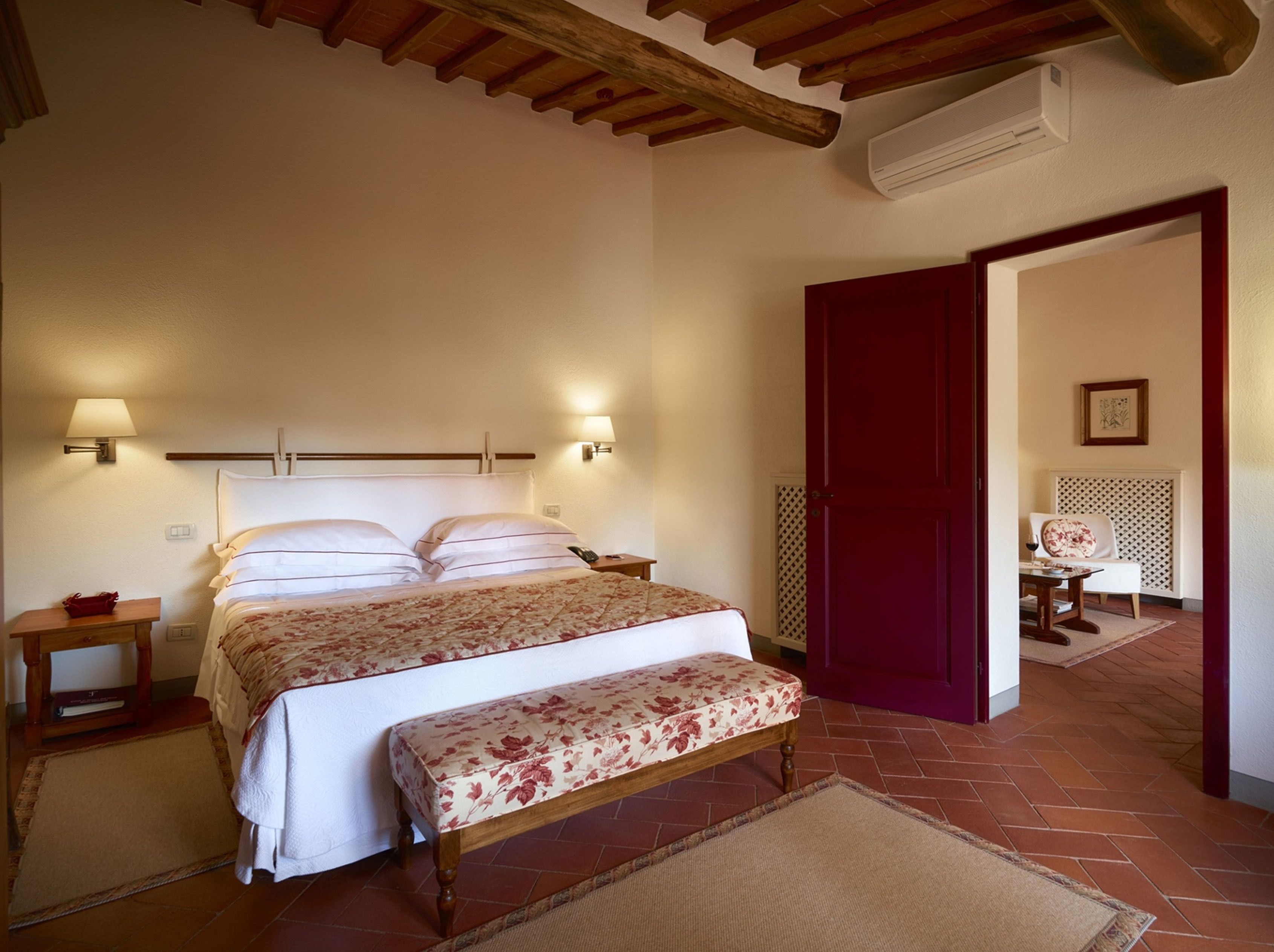 A bedroom with a bed, bedside tables, a bench, and a door leading to another room.