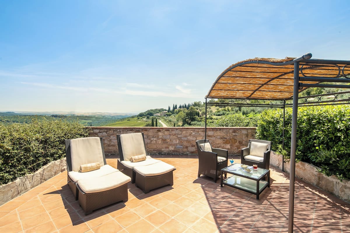 Una terraza con sillones de mimbre, una mesa de vidrio y una pérgola que da sombra.
