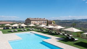 A pool area with lounge chairs and umbrellas, a large building in the background, and a view of the countryside. - Hotel Le Fontanelle (Castelnuovo Berardenga)