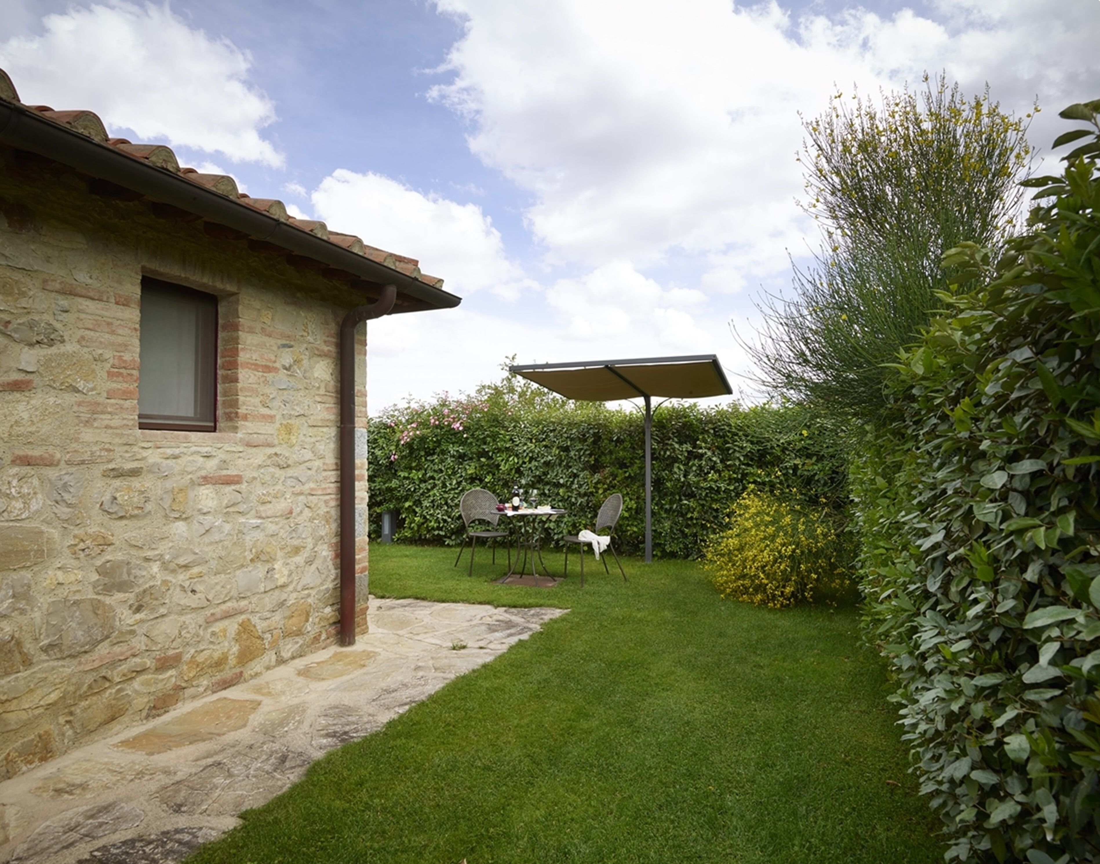 Un bâtiment en pierre avec un toit en tuiles, une cour verdoyante, une table avec des chaises et un espace couvert comprenant une table et des chaises.