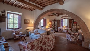 Ampla sala de estar com teto alto, vigas de madeira aparentes e uma grande estrutura de tijolos em arco. Há vários espaços de estar com sofás e poltronas estampadas. Uma lareira e estantes com livros são visíveis ao fundo.