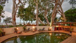 An outdoor swimming pool with lounge chairs and a bar area, surrounded by trees and overlooking the ocean. - Marlin Waters (Palm Cove)