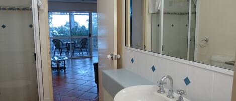 A bathroom with a white sink, a large mirror, and a shower. There is a door leading to a balcony with chairs and a table.