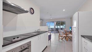 A modern kitchen with a gas stove, oven, and stainless steel range hood. There is a dining area with a table and chairs, and a view of the outdoors through large windows. - Marlin Waters (Palm Cove)
