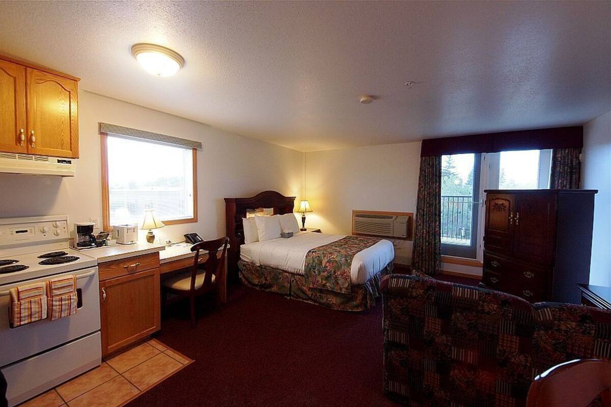 A compact hotel room with a kitchenette, a bed with floral bedding, a chair, and a window with curtains.