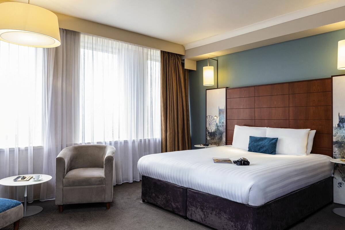 Une chambre d’hôtel avec un grand lit, une chaise, une petite table ronde et une vue sur le paysage urbain par la fenêtre.