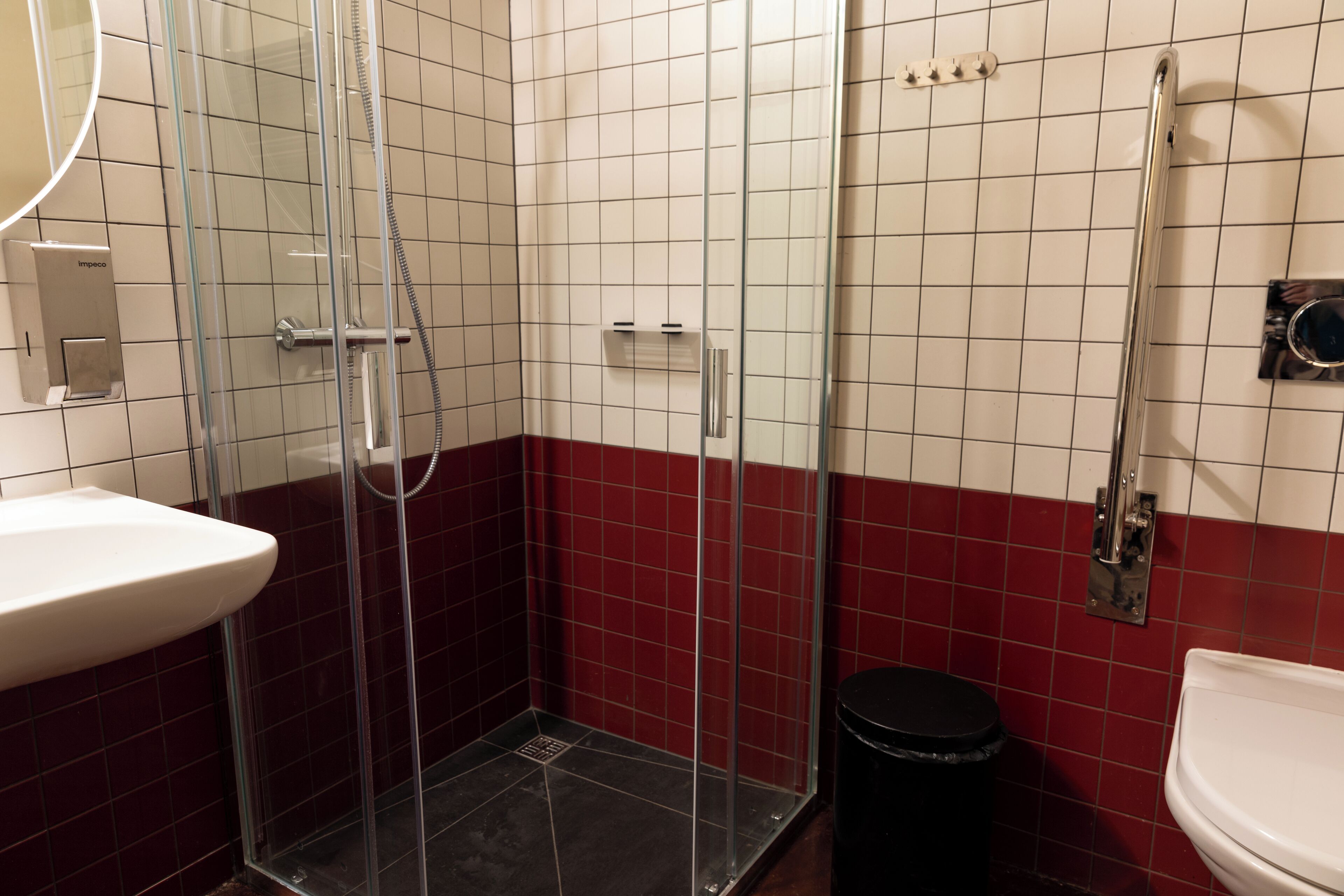Une salle de bain avec une douche à parois de verre, un lavabo blanc et une poubelle noire.