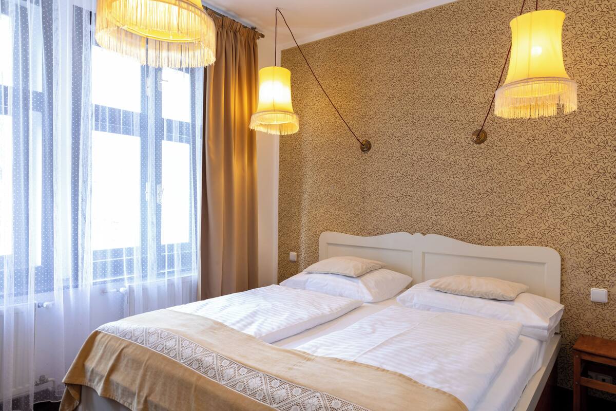 A double bed with a white headboard, two bedside tables, and a window with sheer curtains.