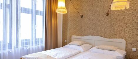 A double bed with a white headboard, two bedside tables, and a window with sheer curtains.