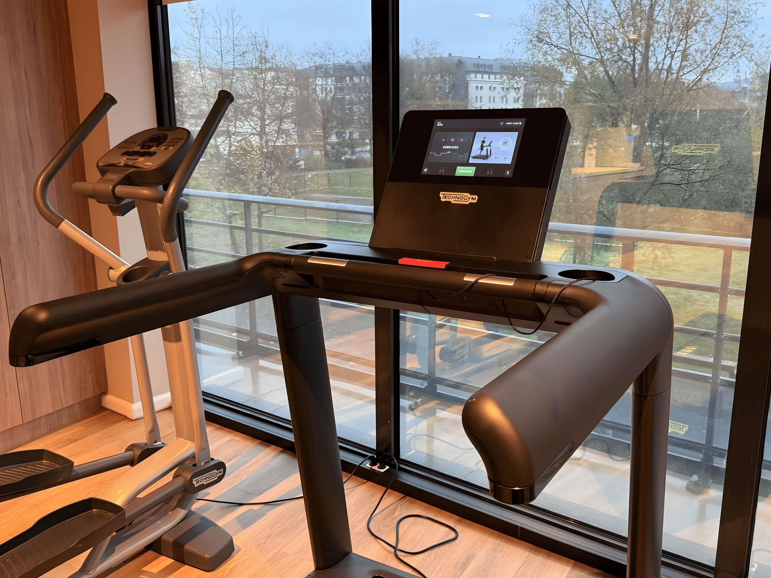 A treadmill with a digital display and a laptop on top, positioned in front of a window with a view of a residential area.